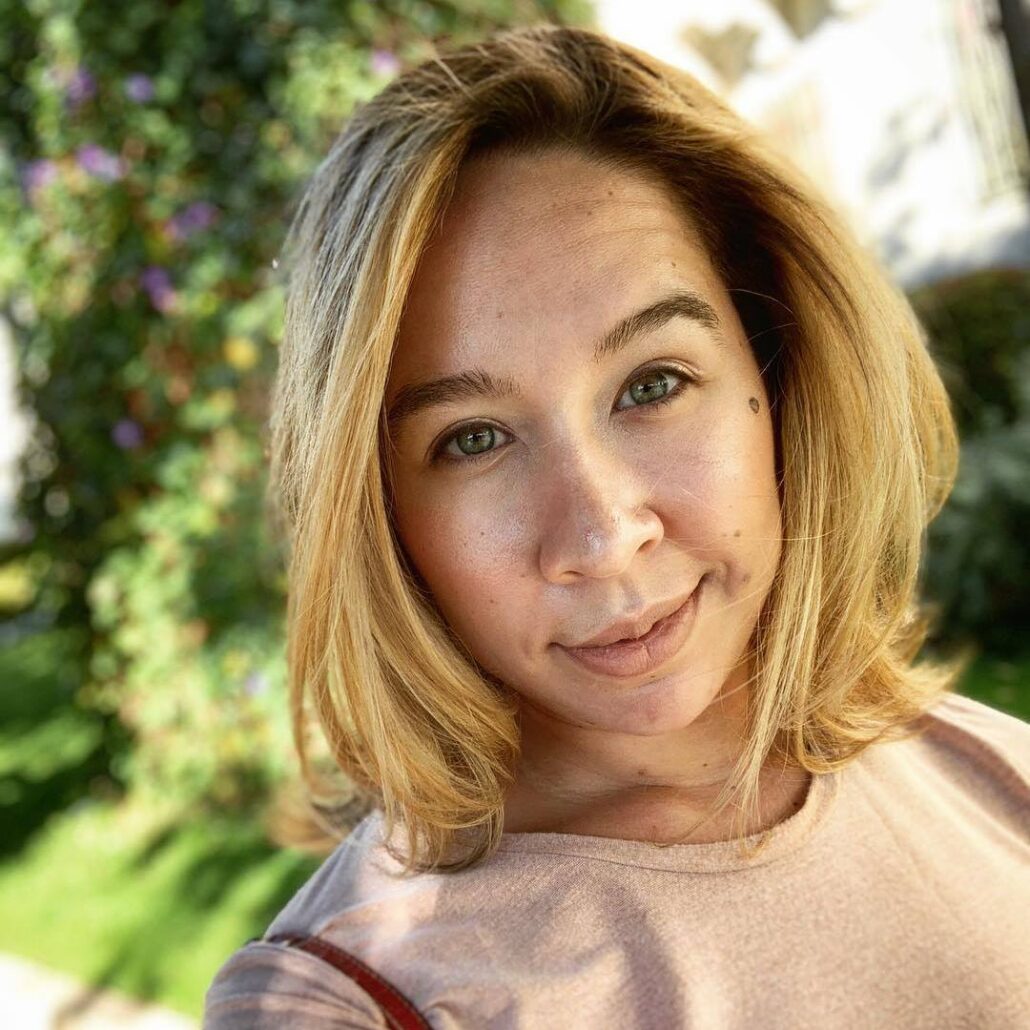Alexa, a woman with blond hair, wears a pale pink t-shirt and stands in front of green foliage.