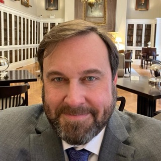 Ed, a white man with brown hair and a beard, wears a white shirt and grey jacket.