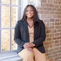 Judge Taylor is a Black woman with dark brown hair. She wears a light tan jumpsuit, a black jacket and a pearl necklace. 