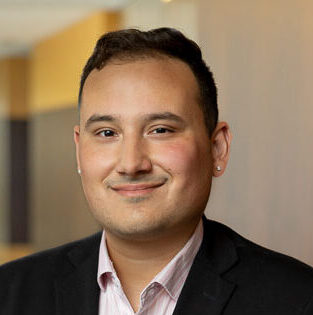 Kevin, a man with dark brown hair, wears a pale pink shirt and a black suit.