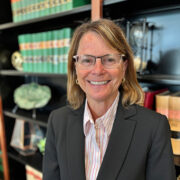 Linda, a white woman with blond hair and glasses, wears a white shirt with pink stripes and dark grey suit.