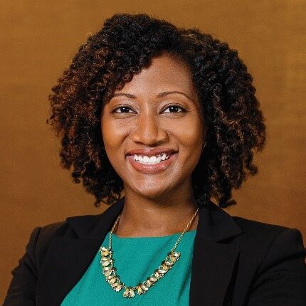 Jocelyn Mallette is a Black woman with brown eyes and curly, shoulder-length hair. She is wearing a teal blouse, gold necklace, and black suit jacket, and she is standing against a warm camel-colored wall.