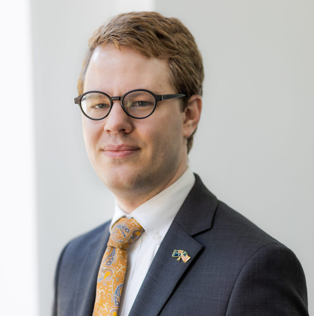 Dan, a white man with brown hair, wears a white shirt, orange tie, and navy jacket.