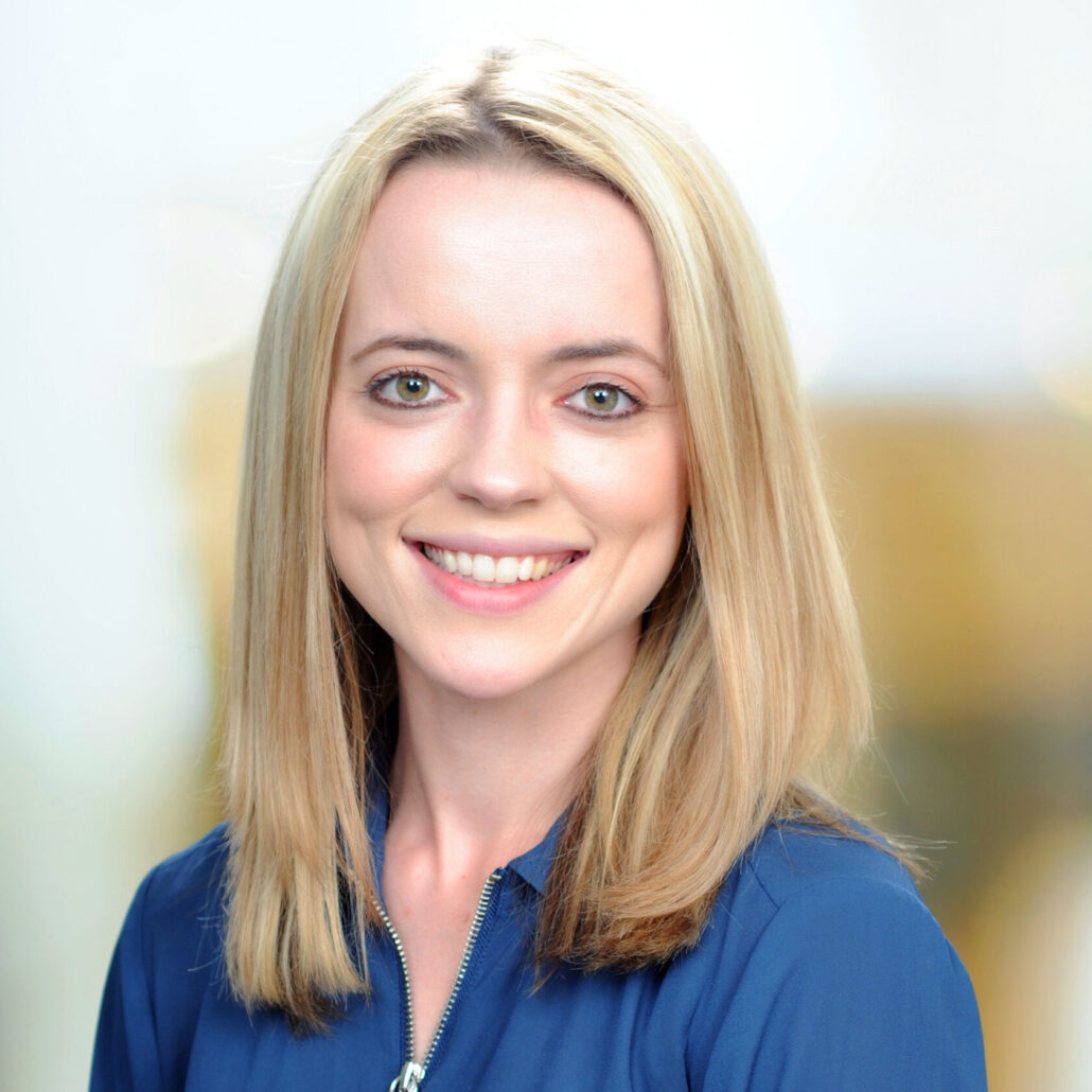 Katie Riddle is a young woman with blond hair and grey eyes. She is pictured smiling and wearing a blue shirt, and she is standing against a light background.
