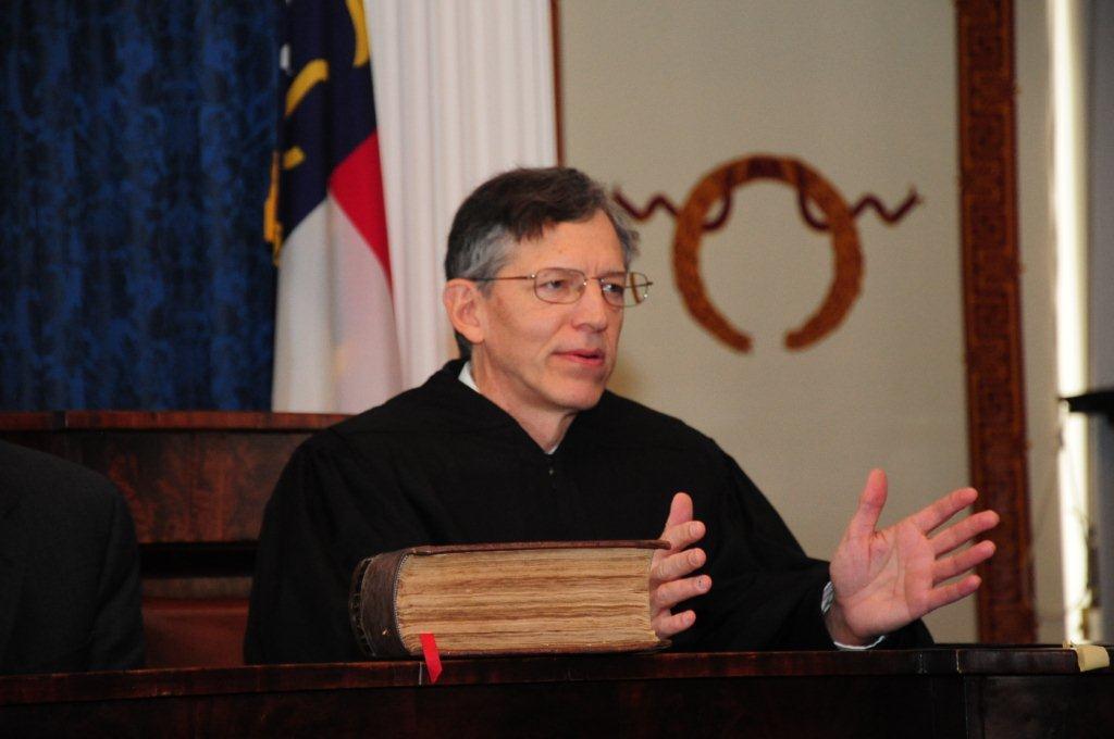 Judge Ward, a man with brown hair, sits on the bench in a courtroom.