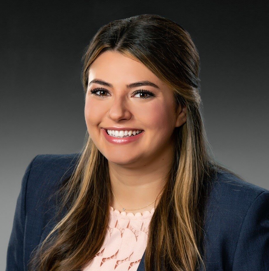 Sarah, a white woman with long golden brown hair, wears a peach blouse and navy suit.