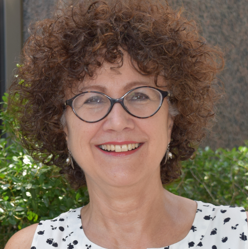 Suzanne, a white woman with curly brown hair, wears a white and blank sleeveless blouse and dark circular glasses. She smiles outside.