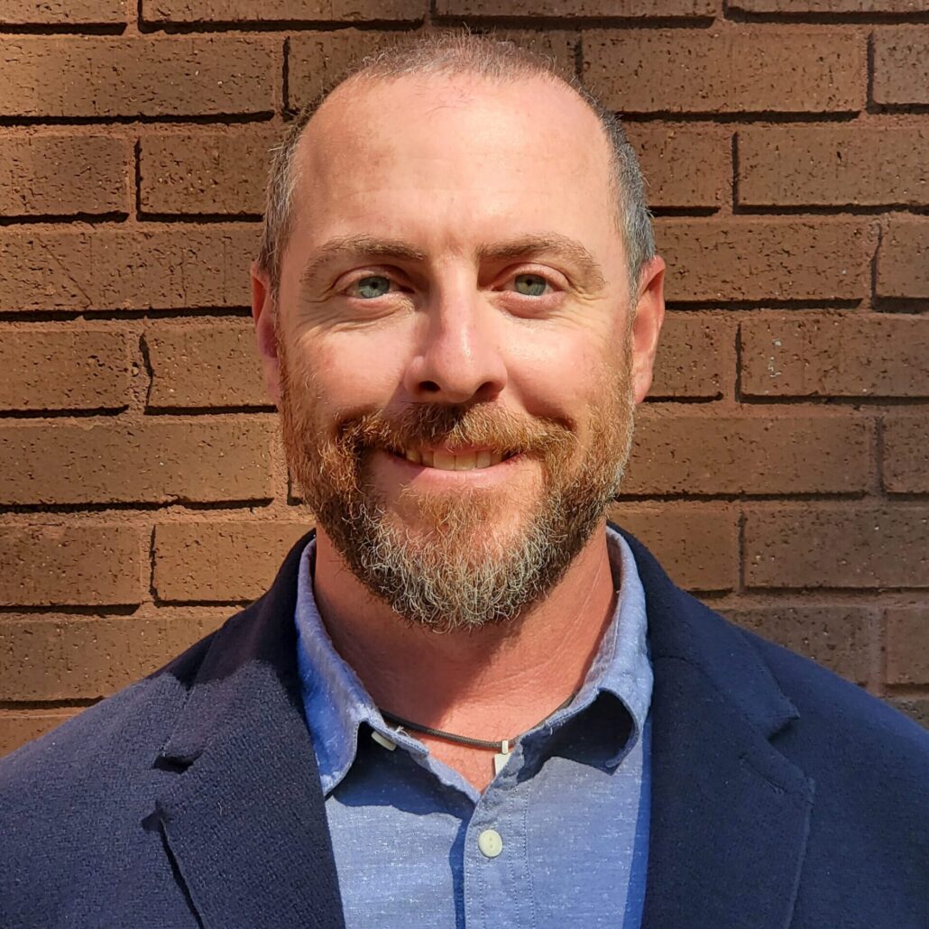 Scott, a white man with a shaved head and a brown beard, wears a blue button-down shirt and navy jacket.