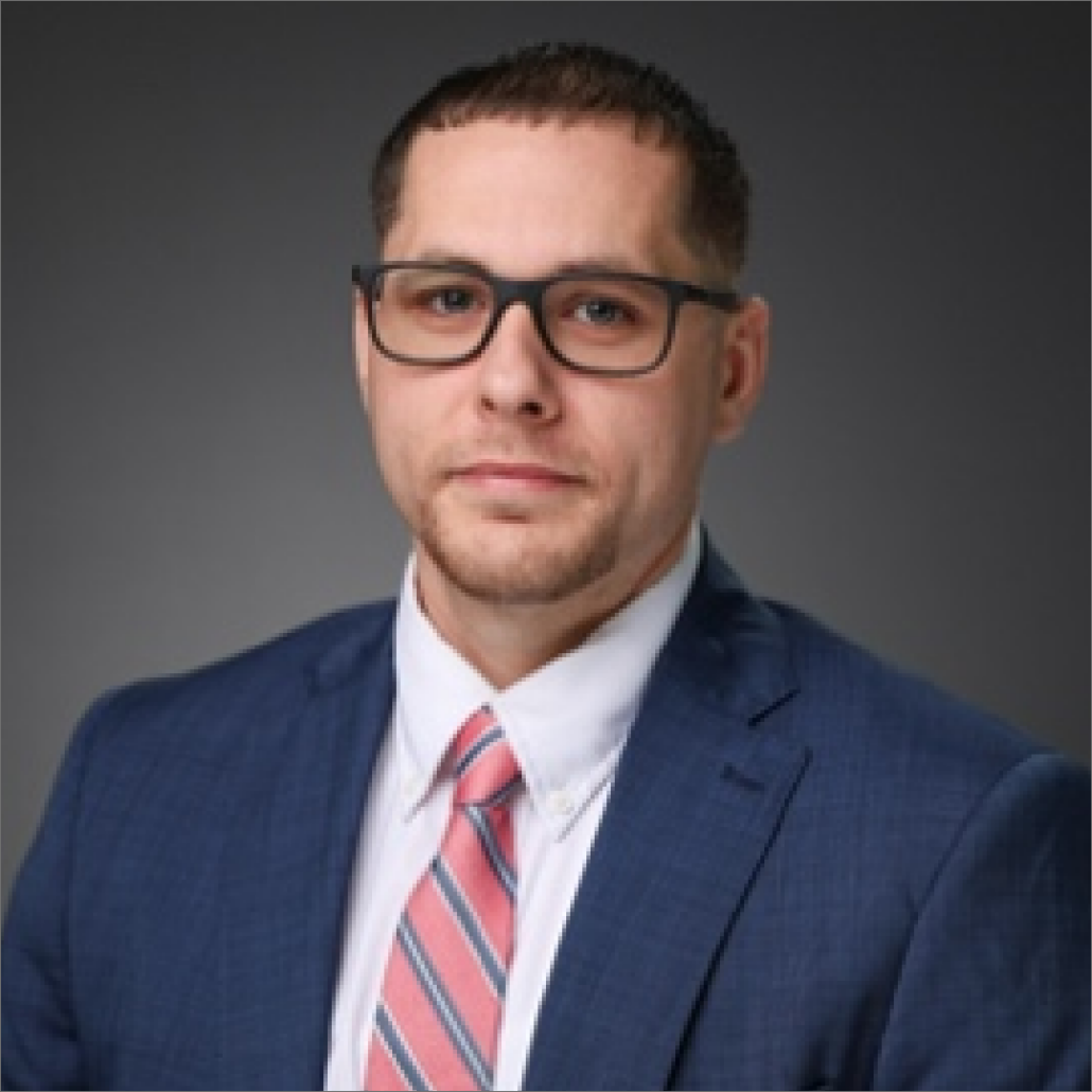 Bryan, a white man with black hair, wears a white shirt, coral and blue striped tie, and blue blazer and black-rimmed glasses.