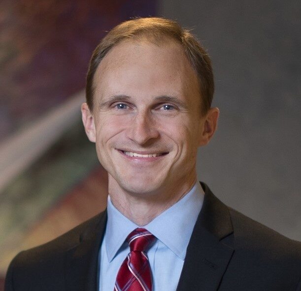 Connor, a white man with brown hair, smiles and is wearing a red tie, pale blue shirt and black blazer.