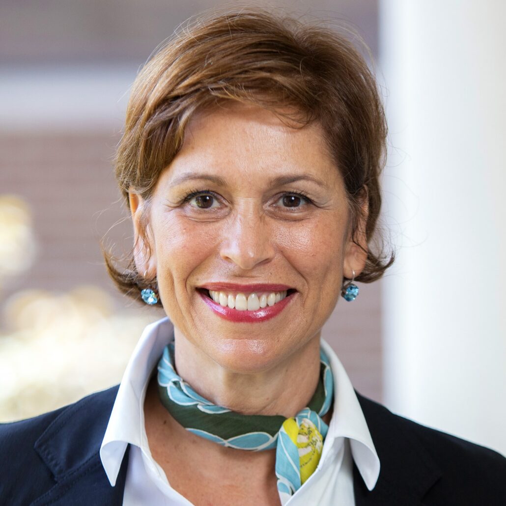 Maria, a woman with short brown hair wears a white shirt, black jacket, and teal, grey, and gold neckerchief.