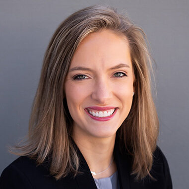 Meredith Parker is a white woman with shoulder-length golden brown hair She is wearing a blue shirt, black jacket and silver necklace, and she is pictured smiling and standing against a grey background.