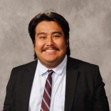 Christian, a man with dark brown hair, wears a white shirt, brown and white striped tie and black suit.