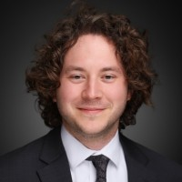 Gabriel, a white man with brown hair, wears a white shirt, black tie and black suit.