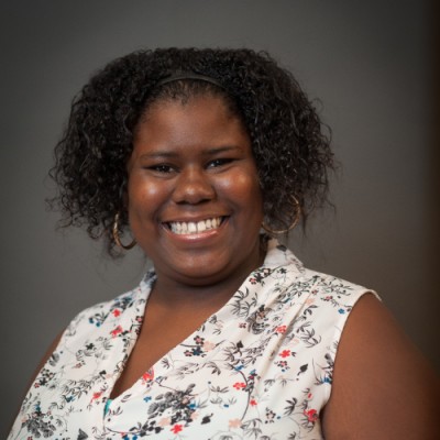 Keyonna, a Black woman with short, curly black hair, wears a white blouse with coral and grey flowers.