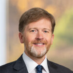 John, a white man with brown hair, wears white shirt, blue tie and dark grey suit.