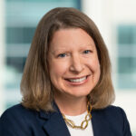 Carolyn, a white woman with brown hair, wears a white blouse, navy suit and gold necklace.