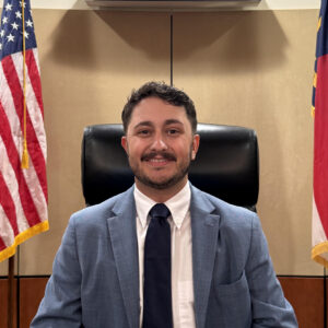 Steven, a white man with brown hair, wears a white shirt, black tie and light grey suit. The American flag and North Carolina flag are behind him.