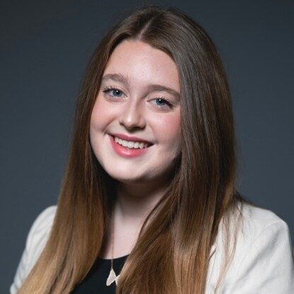 Sydney, a white woman with brow hair, wears a black blouse and a cream-colored jacket.