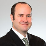 James, a white man with brown hair, wears a white shirt, green plaid tie and black suit.