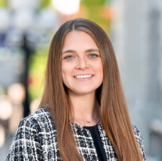 Hannah, a white woman with brown hair wears a black blouse and plaid suit.