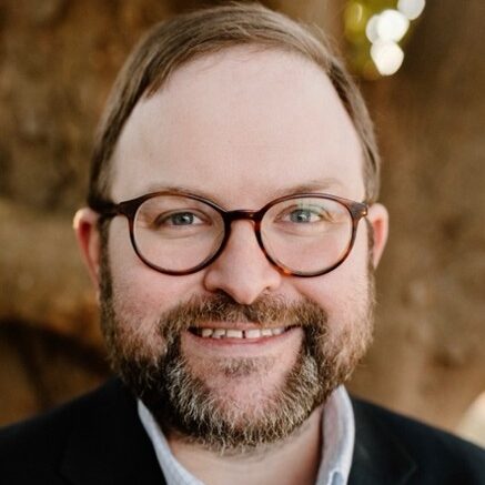 Jason, a white man with brown hair and a beard, wears brown glasses, a white shirt and black jacket.