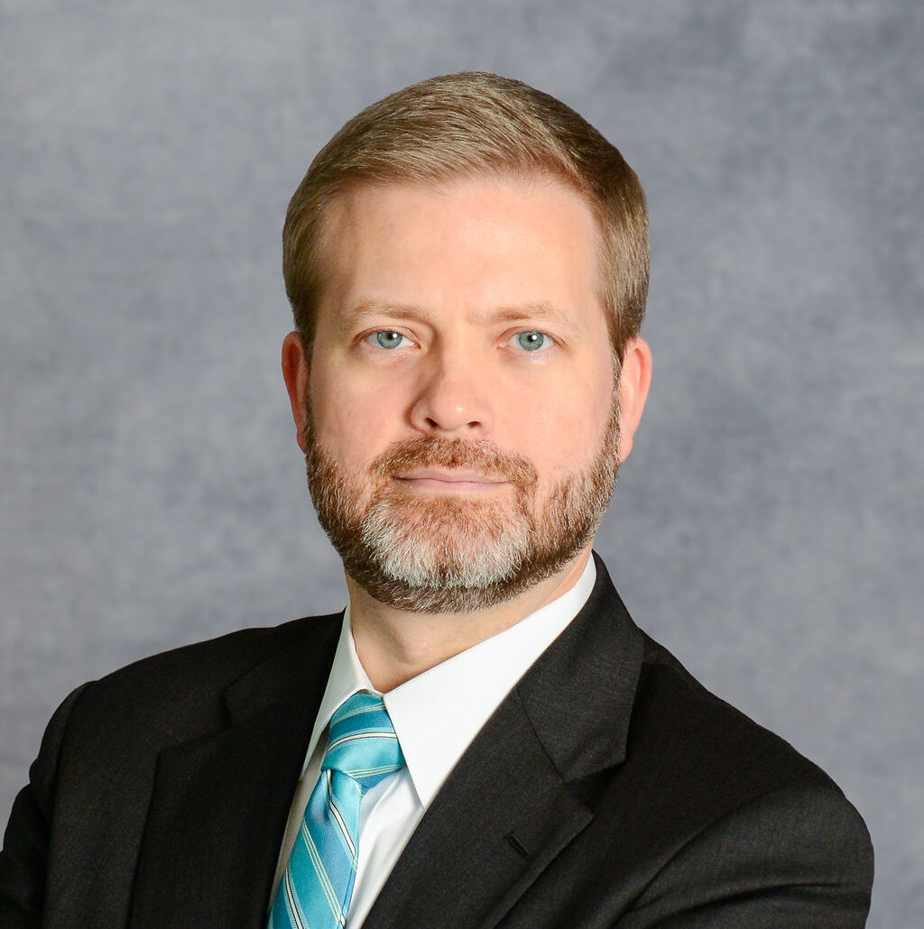 Isaac, a white man with brown hair and a beard, wears a white shirt, teal tie and black suit.