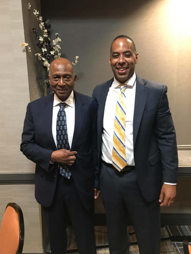 Smith is a Black man with black hair. He wears a white shirt, navy tie and black suit. Davis is a Black man with black hair. He wears a white shirt, yellow tie and black suit. 