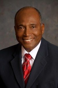 Smith, a Black man with black hair, wears a white shirt, red tie and black suit.