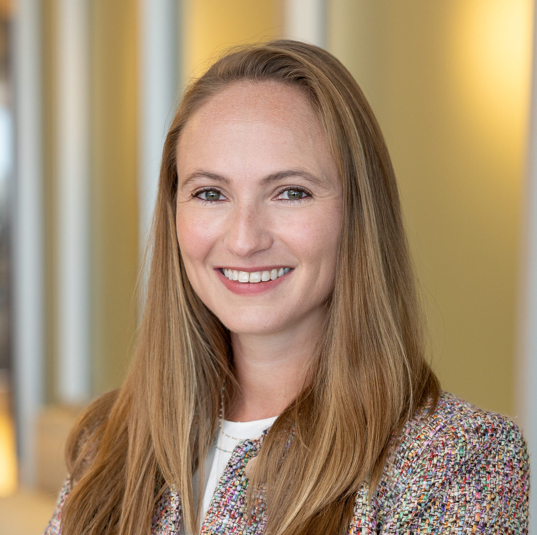 Ella, a white woman with light brown hair, wears a white blouse and multicolored button down short-sleeve blazer.