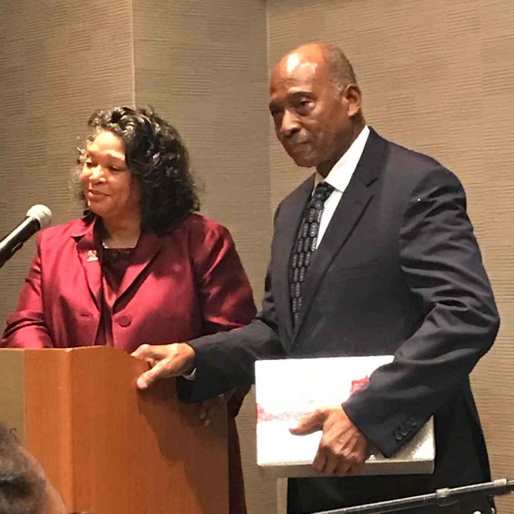 Smith, a Black man, wears a white shirt, dark tie and black suit. Taylor is a Black woman with black curly hair, and she is wearing a burgundy suit.
