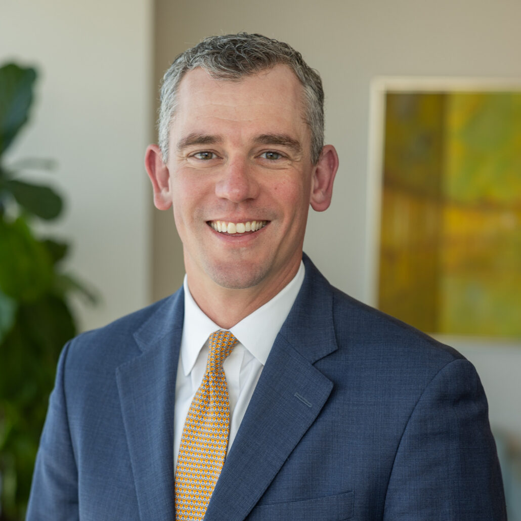 Chad, a white man with brown and grey hair, wears a white shirt, yellow tie and grey suit.